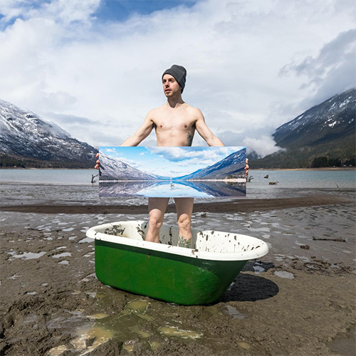 Photo of Jeremie standing in a bathtub holding a piece of art, in front of a mountain backdrop