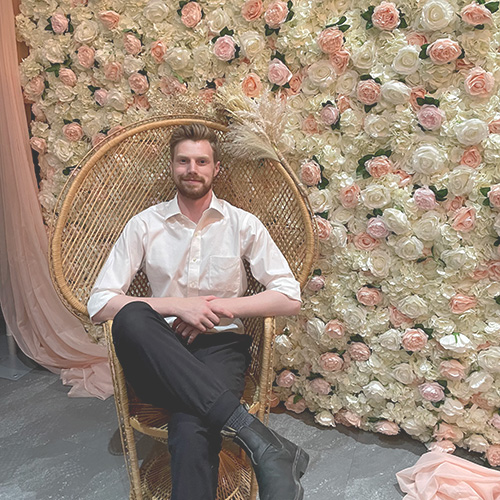 Photo of Ryan Schmidt Sitting in Front of a Wall of Flowers
